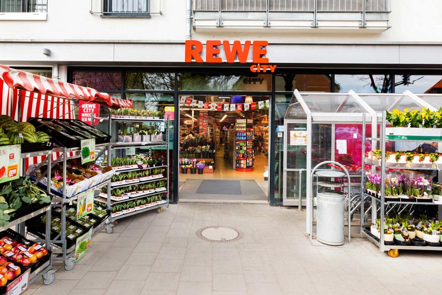 Eingang eines REWE City Markts mit Obst- und Pflanzenständen.